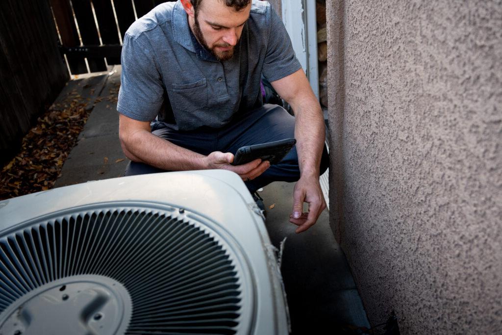Young Male Property Inspector Photographing a Line to a Residential Air Conditioner Condenser Unit at the side of a stucco home